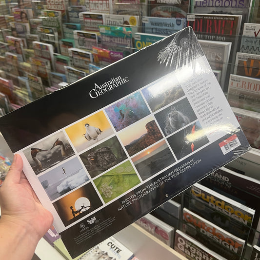A hand holds a glossy, shrink-wrapped Australian Geographic magazine in a retail store setting with shelves of magazines in the background.