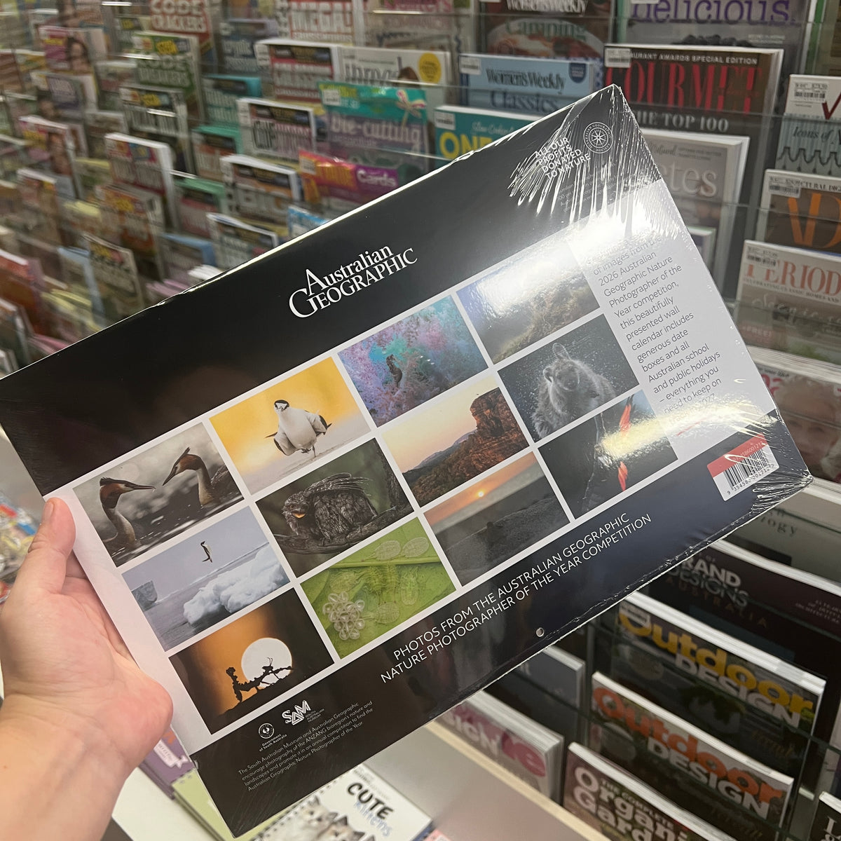 A hand holds a glossy, shrink-wrapped Australian Geographic magazine in a retail store setting with shelves of magazines in the background.