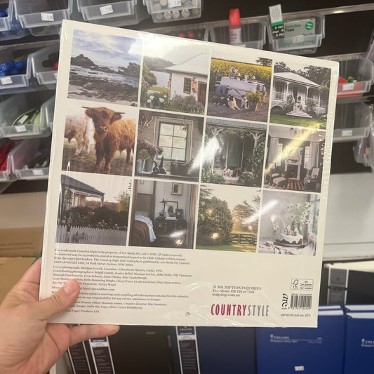 A hand holds a shrink-wrapped photography album or book titled 'COUNTRYSTYLE' in a retail storage area. The back cover features a grid of outdoor and interior lifestyle photographs, product text, and a barcode.