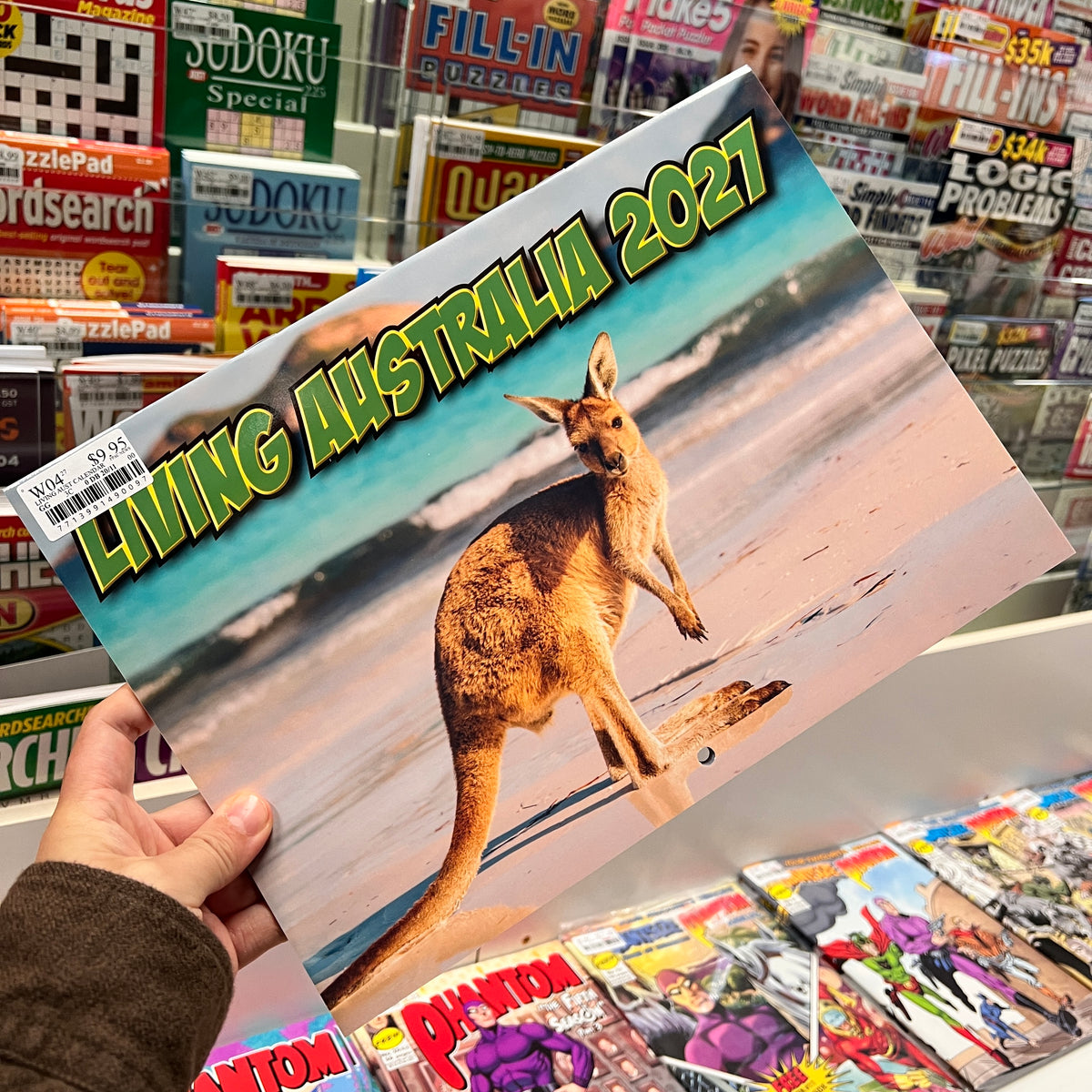 A hand holds a 2027 calendar titled 'LIVING AUSTRALIA 2027' featuring a kangaroo on a beach, with a background of a book and magazine store.