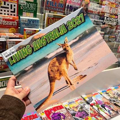 A hand holds a 2027 calendar titled 'LIVING AUSTRALIA 2027' featuring a kangaroo on a beach, with a background of a book and magazine store.