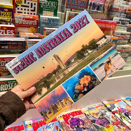 A hand holds a 2027 Scenic Australia calendar in a retail store setting, with shelves of books and magazines in the background.