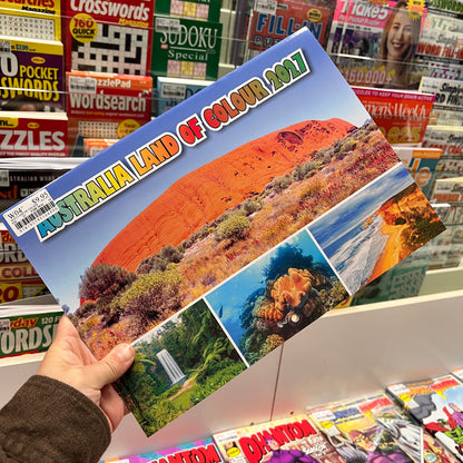A hand holds a printed postcard or small print featuring Australia Land of Colour 2021, with a large red rock formation and smaller scenic panels on the front. The background shows a magazine or book display shelf with colorful covers.