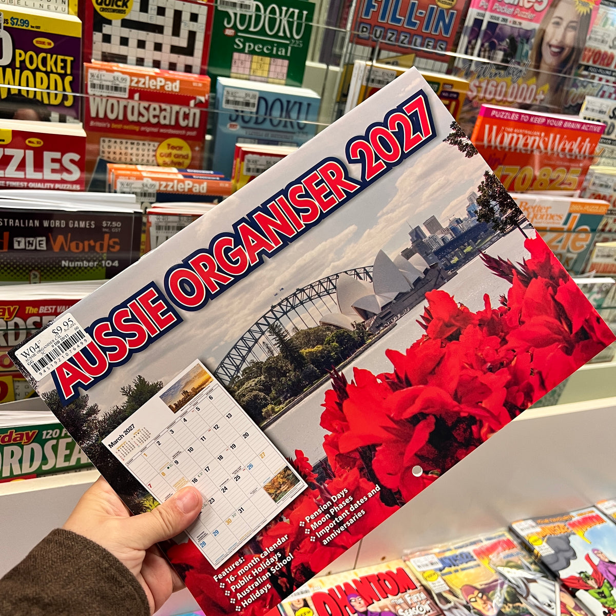 A hand holds a 2027 Aussie Organiser desk calendar in a retail store. The calendar cover features a Sydney Opera House, Sydney Harbour Bridge, and bright red flowers, with a small monthly calendar preview visible on the lower left.
