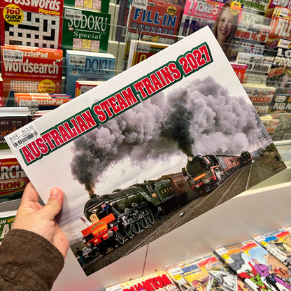 A hand holds a printed magazine or booklet titled "Australian Steam Trains 2021" featuring a steam locomotive scene, with a retail shelf of books and magazines in the background.