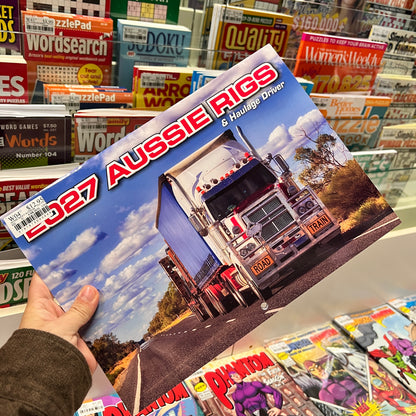 A hand holds a copy of the 'CD27 Aussie Rigs & Haulage Driver' magazine or book in front of a store shelf filled with other books and magazines.