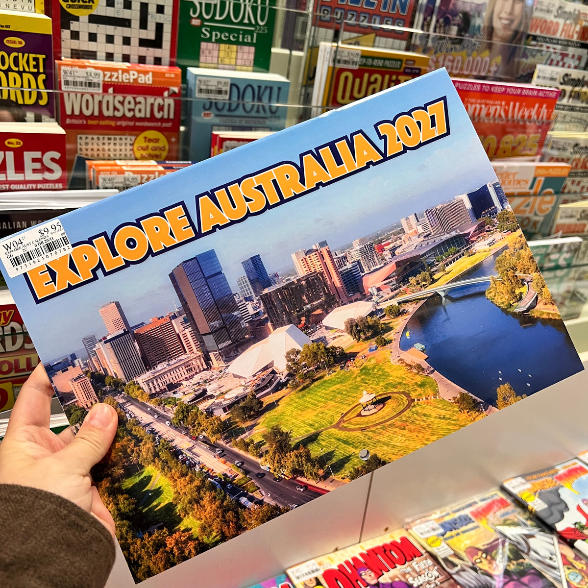 A hand holds a printed poster or booklet titled "EXPLORE AUSTRALIA 2027" with a cityscape and park scene on the cover, photographed in a retail store aisle with Sudoku and other magazines in the background.