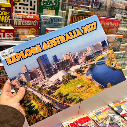 A hand holds a printed poster or booklet titled "EXPLORE AUSTRALIA 2027" with a cityscape and park scene on the cover, photographed in a retail store aisle with Sudoku and other magazines in the background.