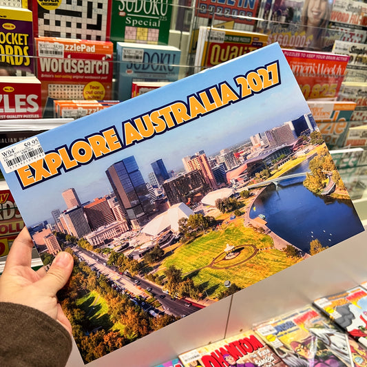 A hand holds a printed poster or booklet titled "EXPLORE AUSTRALIA 2027" with a cityscape and park scene on the cover, photographed in a retail store aisle with Sudoku and other magazines in the background.