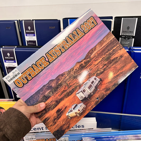 A hand holds a Collins Vanesa 'Outback Australia 2021' pocket program or booklet in a retail store setting, with blue binders in the background.