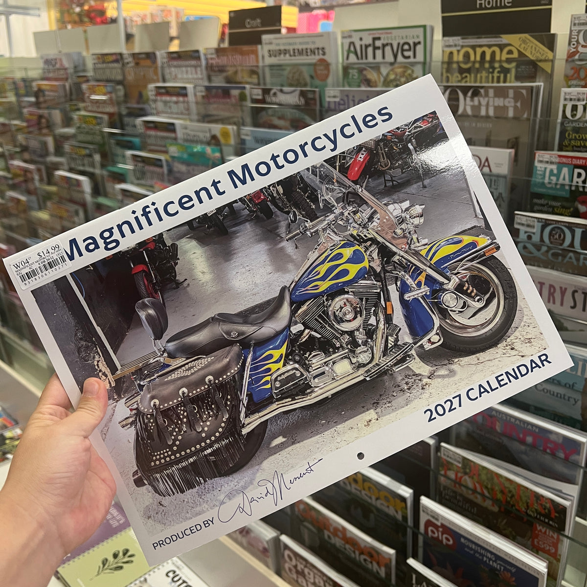 A hand holds a 2027 calendar titled 'Magnificent Motorcycles' in a retail store. The calendar cover features a photograph of a blue and yellow flame-patterned motorcycle, with the text '2027 CALENDAR' in the bottom right corner.