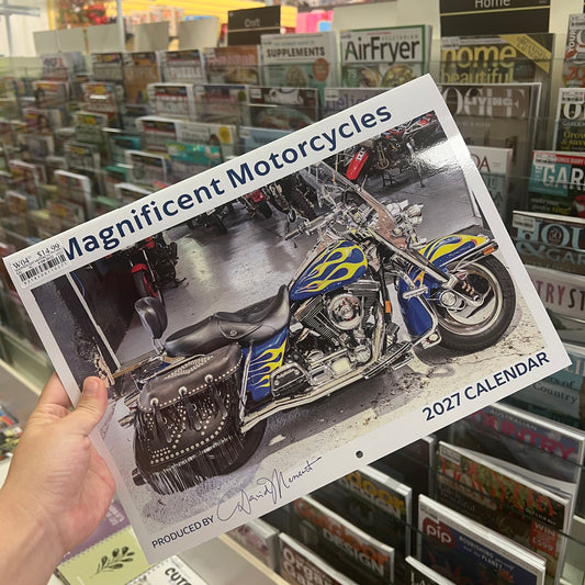 A hand holds a 2027 calendar titled 'Magnificent Motorcycles' in a retail store. The calendar cover features a photograph of a blue and yellow flame-patterned motorcycle, with the text '2027 CALENDAR' in the bottom right corner.