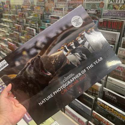 A hand holds a glossy 2027 Nature Photographer of the Year calendar featuring penguins and a seal on the cover, with a store shelf of magazines in the background.