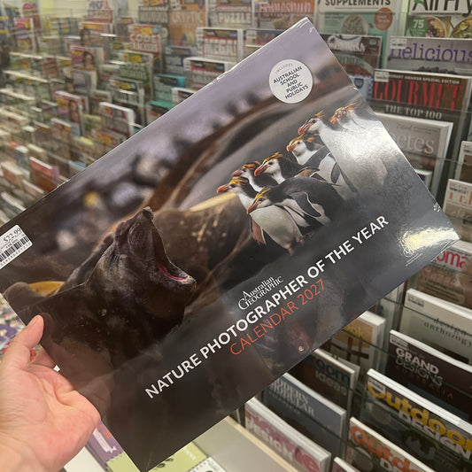 A hand holds a glossy 2027 Nature Photographer of the Year calendar featuring penguins and a seal on the cover, with a store shelf of magazines in the background.