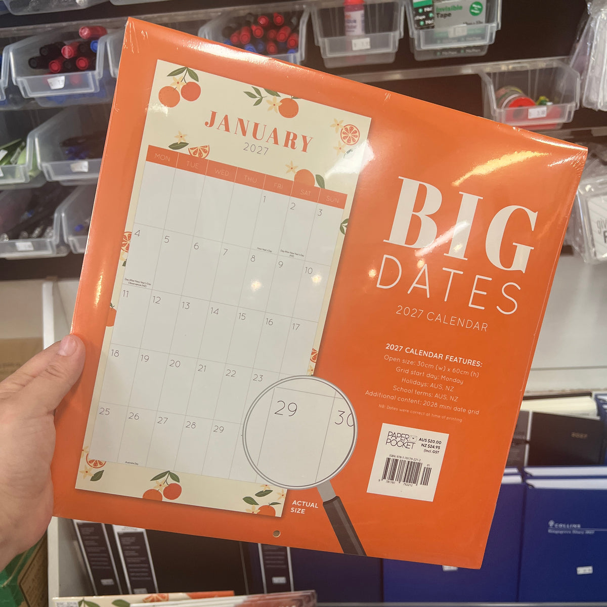 A hand holds a shrink-wrapped 2027 calendar titled "BIG DATES" in orange. The calendar cover features a monthly calendar preview for January 2027, orange fruit illustrations, and a magnifying glass graphic.