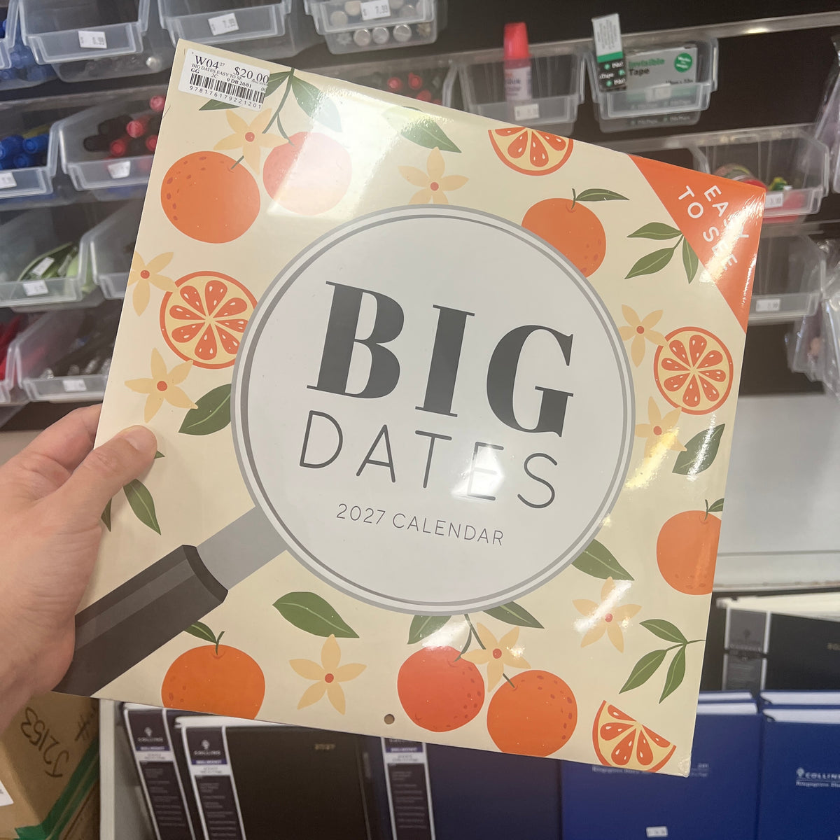 A hand holds a 2027 calendar titled 'BIG DATES' with an orange and cream pattern, in a retail store setting.