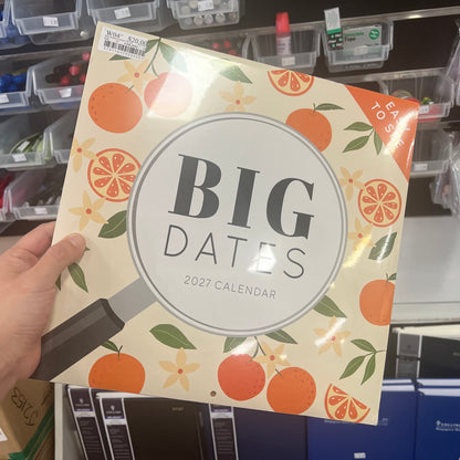 A hand holds a 2027 calendar titled 'BIG DATES' with an orange and cream pattern, in a retail store setting.
