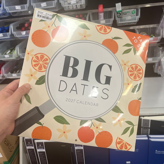 A hand holds a 2027 calendar titled 'BIG DATES' with an orange and cream pattern, in a retail store setting.