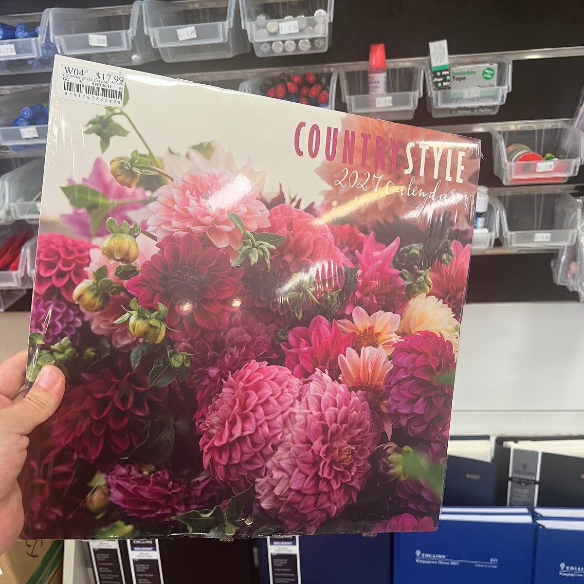 A hand holds a 2027 'Countryside' annual calendar featuring a vibrant cover of pink, red, and purple dahlias. The calendar is photographed in a retail store setting with shelves of plastic bins and products in the background.