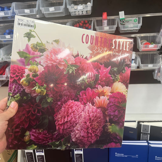 A hand holds a 2027 'Countryside' annual calendar featuring a vibrant cover of pink, red, and purple dahlias. The calendar is photographed in a retail store setting with shelves of plastic bins and products in the background.