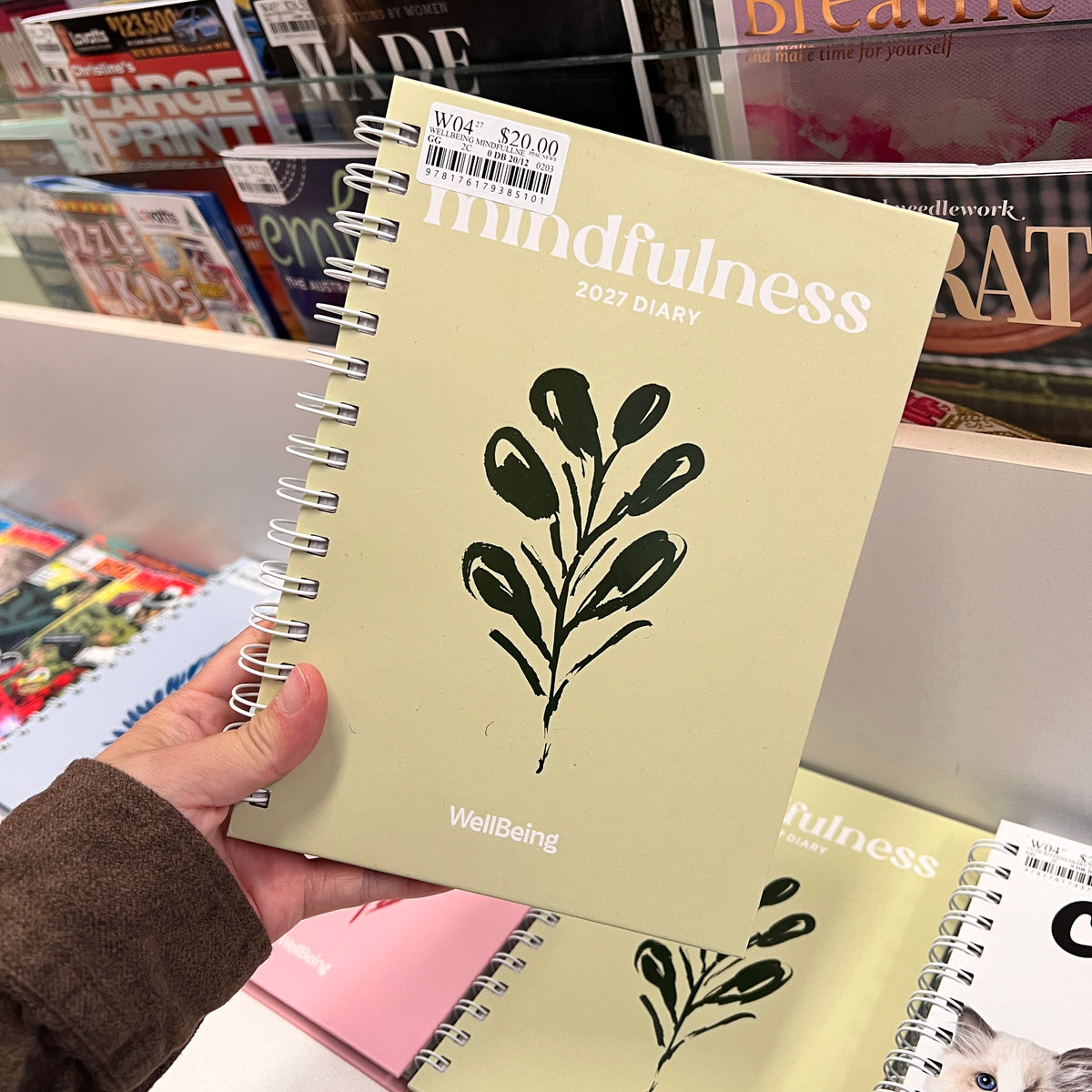 A hand holding a pale yellow 2027 spiral-bound diary titled 'mindfulness' with a black botanical leaf illustration on the cover,