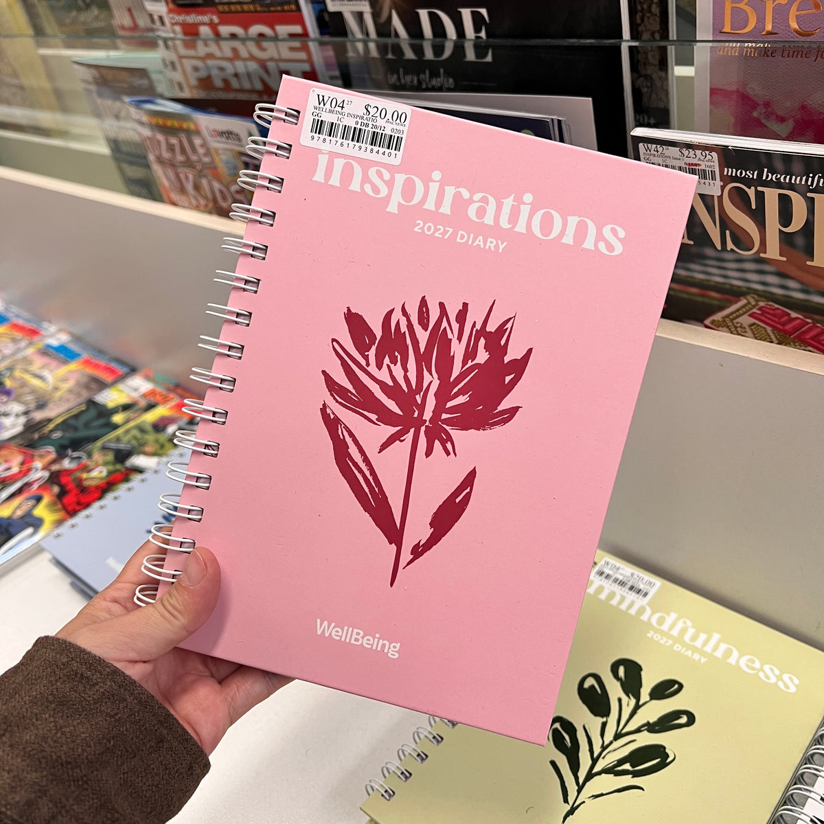 A hand holds a pink spiral-bound 2023 diary titled 'inspirations' with a red floral graphic and 'WellBeing' printed at the bottom. The background shows a retail bookshelf with other books and magazines.