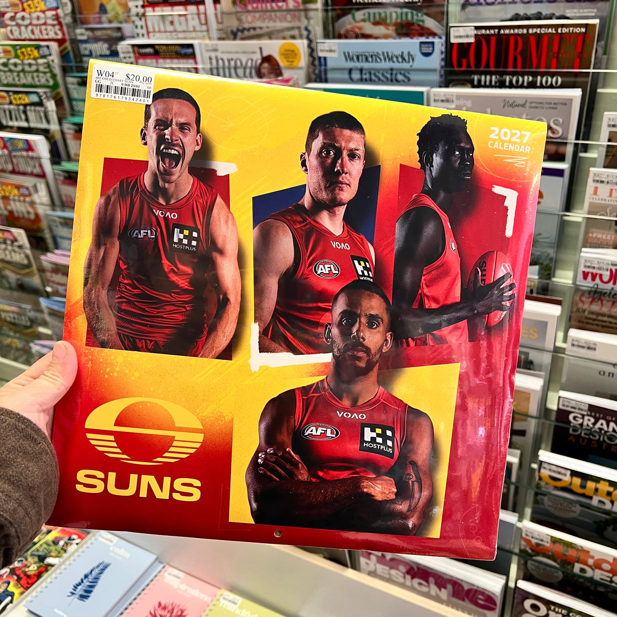A hand holds a 2027 AFL calendar featuring the Suns brand and photos of three football players in red jerseys, set against a background of a book and magazine store.