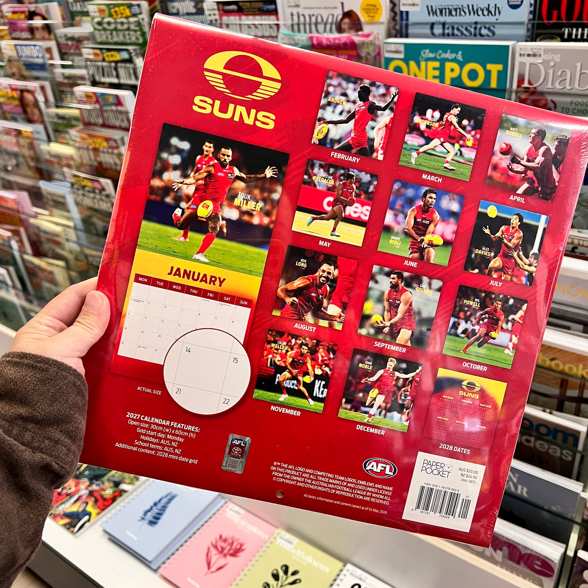 A hand holds a red AFL calendar featuring the Suns brand, with monthly photo spreads and a January calendar page visible, photographed in a retail store setting.
