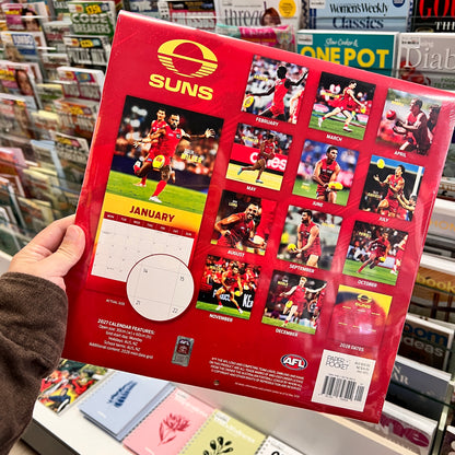 A hand holds a red AFL calendar featuring the Suns brand, with monthly photo spreads and a January calendar page visible, photographed in a retail store setting.