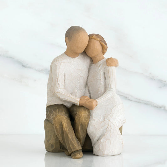 A willow tree figurine of a seated couple embracing, with a man on the left in a textured white shirt and brown pants and a woman on the right in a white dress. The figures have smooth, matte, sculpted surfaces.