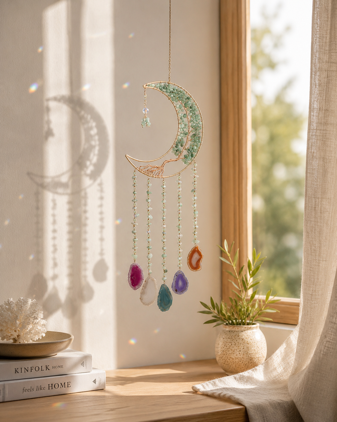 A decorative crescent moon suncatcher with hanging colorful beads is displayed on a windowsill in a warm, sunlit room with a small potted plant and books nearby.