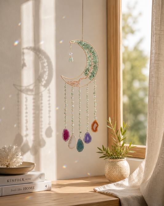 A decorative crescent moon suncatcher with hanging colorful beads is displayed on a windowsill in a warm, sunlit room with a small potted plant and books nearby.