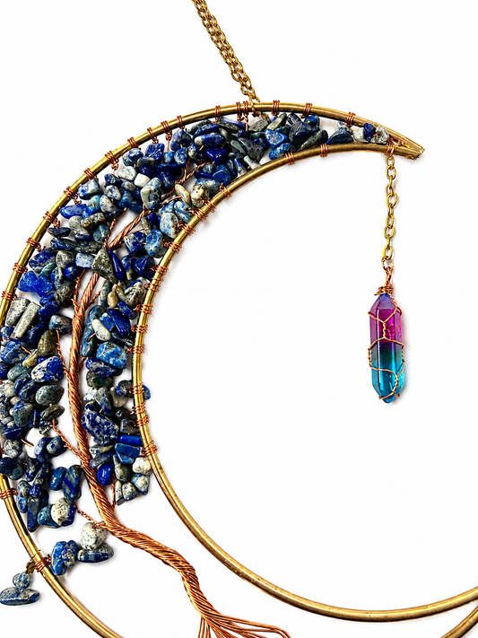 A crescent moon-shaped gold wire dream catcher adorned with blue and white lapis lazuli crystal chips, featuring a dangling magenta and blue gradient crystal pendant on a white background.