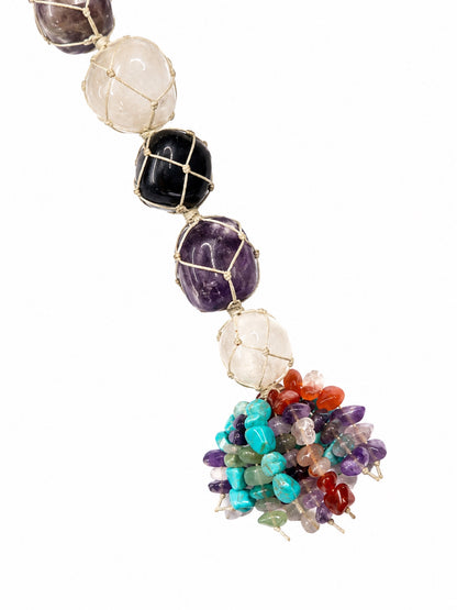 A beaded hanging pendant featuring large faceted gemstone beads in white, black, and purple, suspended in gold wire mesh, with a colorful tassel of small multicolored crystal chips at the bottom, shown against a plain white background.