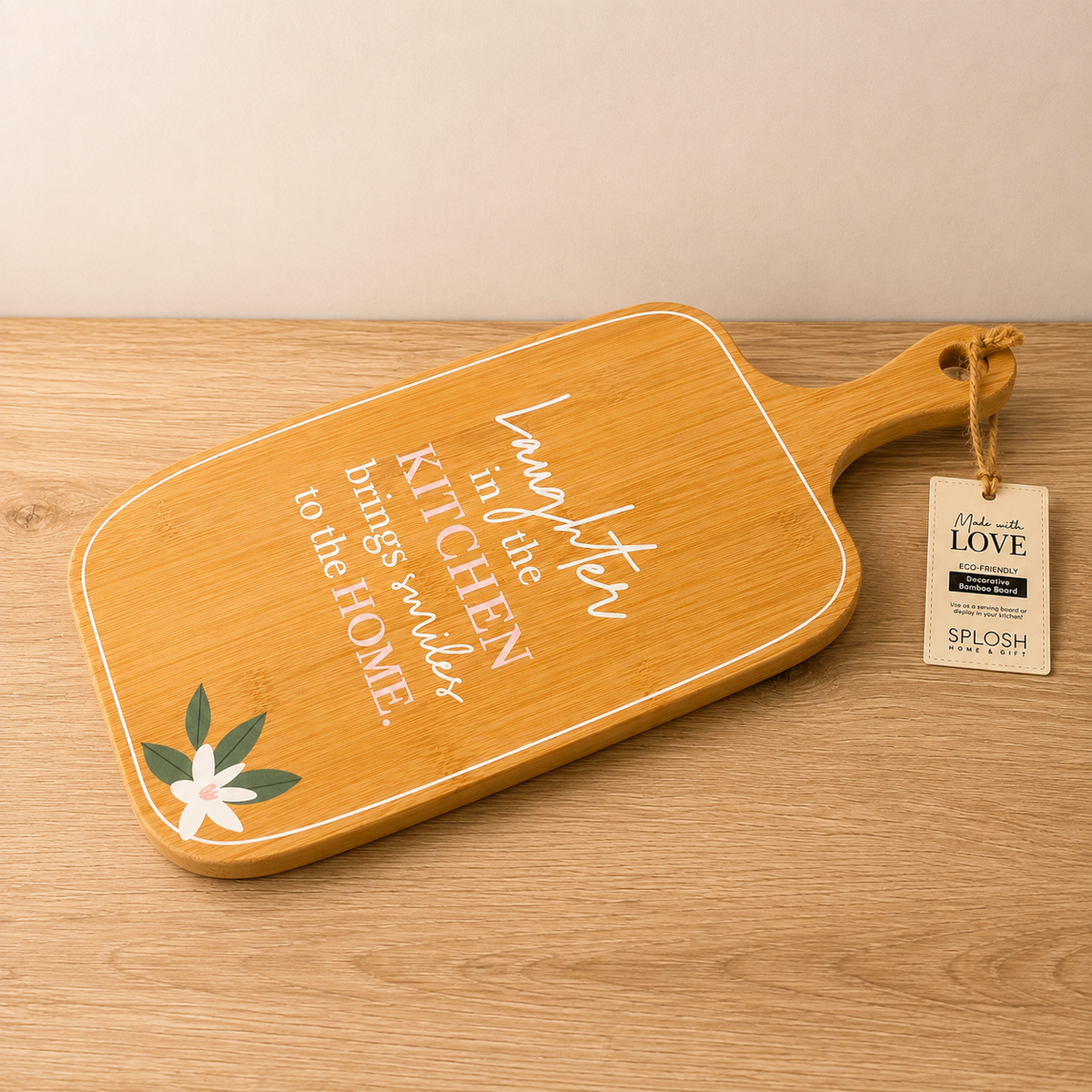 A rectangular wooden cutting board with a handle, featuring white script text that reads 'Laughter in the kitchen brings smiles to the home' and a small white flower with green leaves in the lower left corner. 