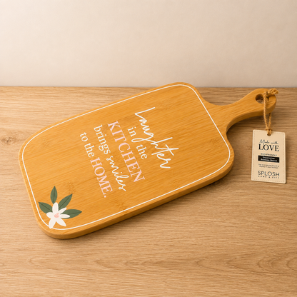 A rectangular wooden cutting board with a handle, featuring white script text that reads 'Laughter in the kitchen brings smiles to the home' and a small white flower with green leaves in the lower left corner. 