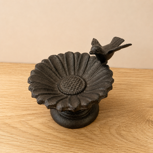 A dark cast iron bird feeder shaped like a sunflower with a small bird perched on the side, displayed on a light wood surface against a beige background.