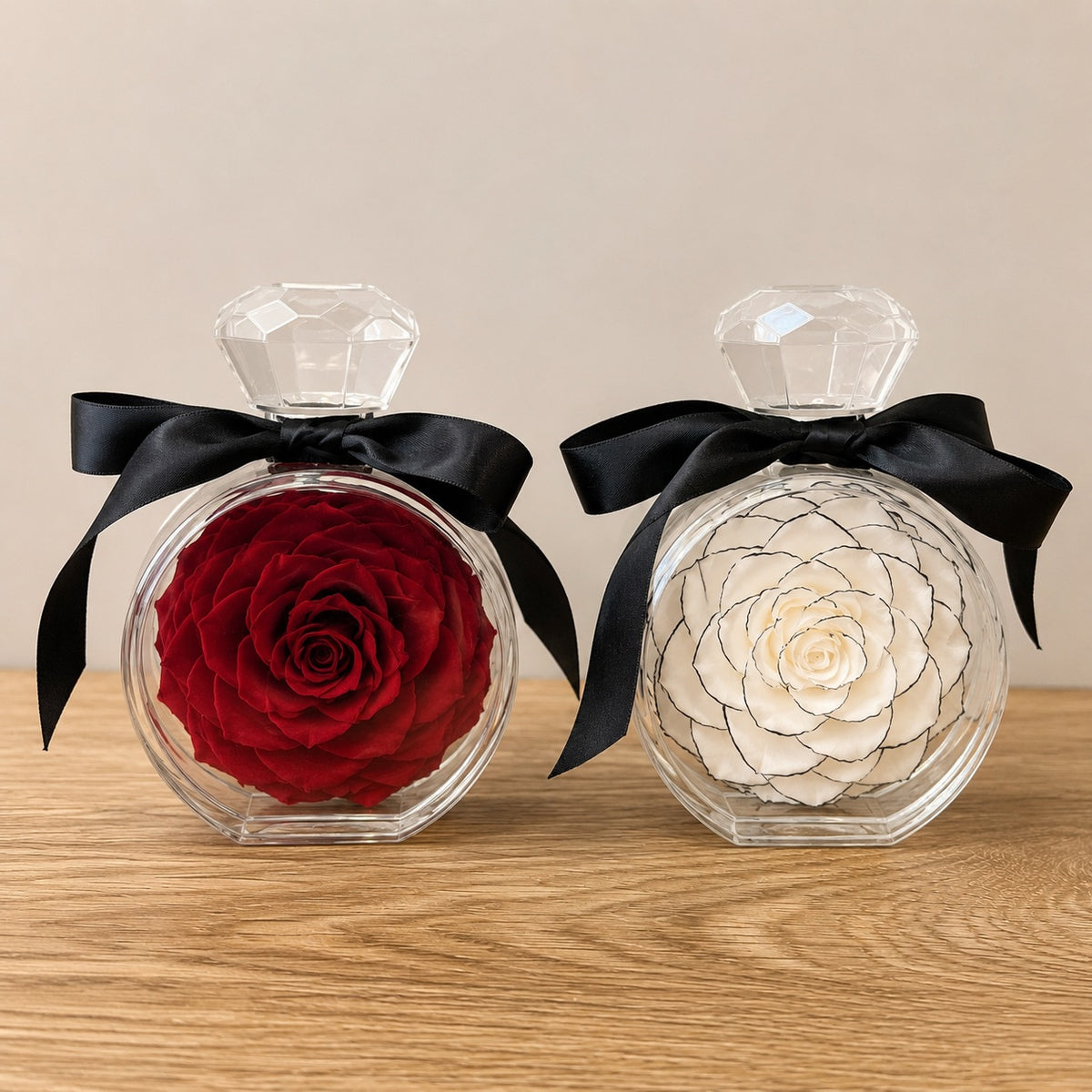 Two round clear perfume bottles with faceted crystal-style caps and black satin bows, displayed on a wooden surface against a beige wall. One bottle contains a preserved red rose, and the other contains a preserved off-white rose with dark linework details.
