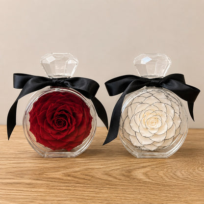 Two round clear perfume bottles with faceted crystal-style caps and black satin bows, displayed on a wooden surface against a beige wall. One bottle contains a preserved red rose, and the other contains a preserved off-white rose with dark linework details.