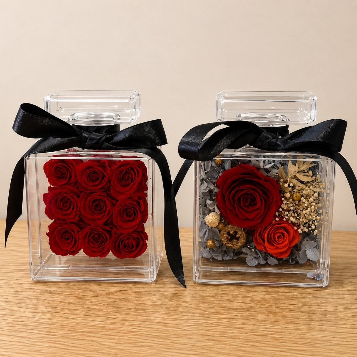 Two clear square flower box, tied with black ribbons, displayed on a light wood surface against a beige wall. The left box contains nine preserved red roses, and the right box contains one large red rose, a smaller red rose, dried gold filler, and gray foliage.