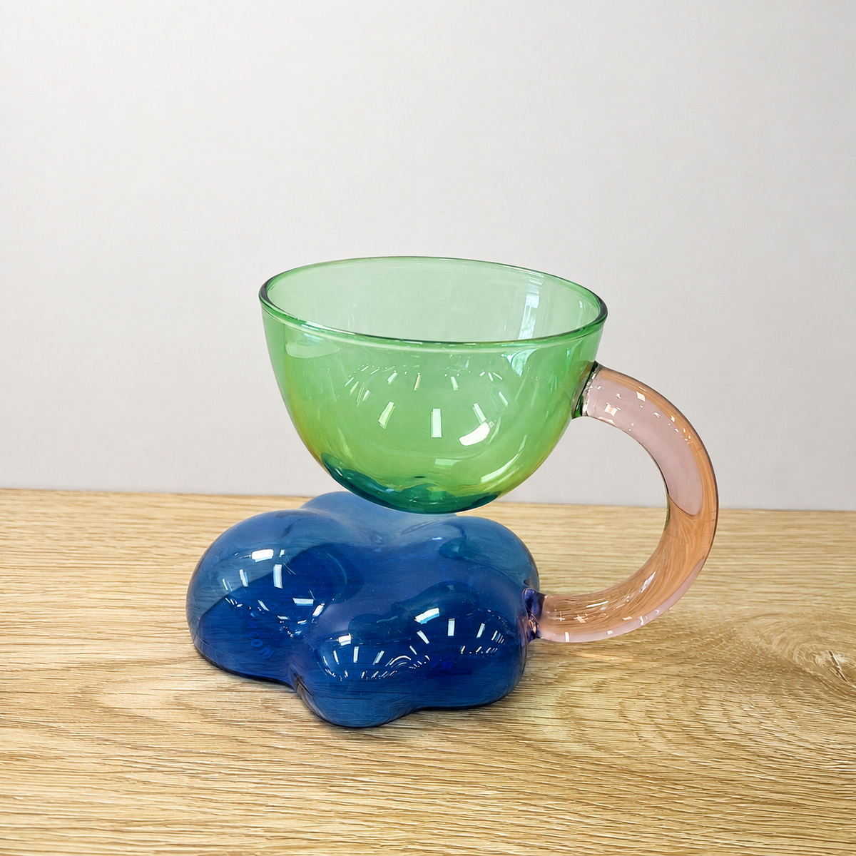 A translucent glass mug with a cloud-shaped blue base, a green bowl-shaped top, and a pink handle, shown on a light wood tabletop against a plain light gray wall.