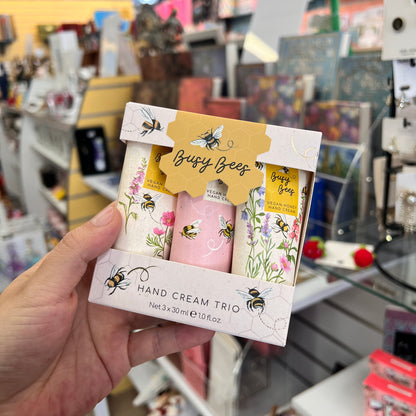 A hand holds a boxed hand cream trio labeled 'Busy Bees' in a store setting. The packaging is white with yellow and green floral/bee illustrations, and the three cream tubes inside are white, pink, and yellow.