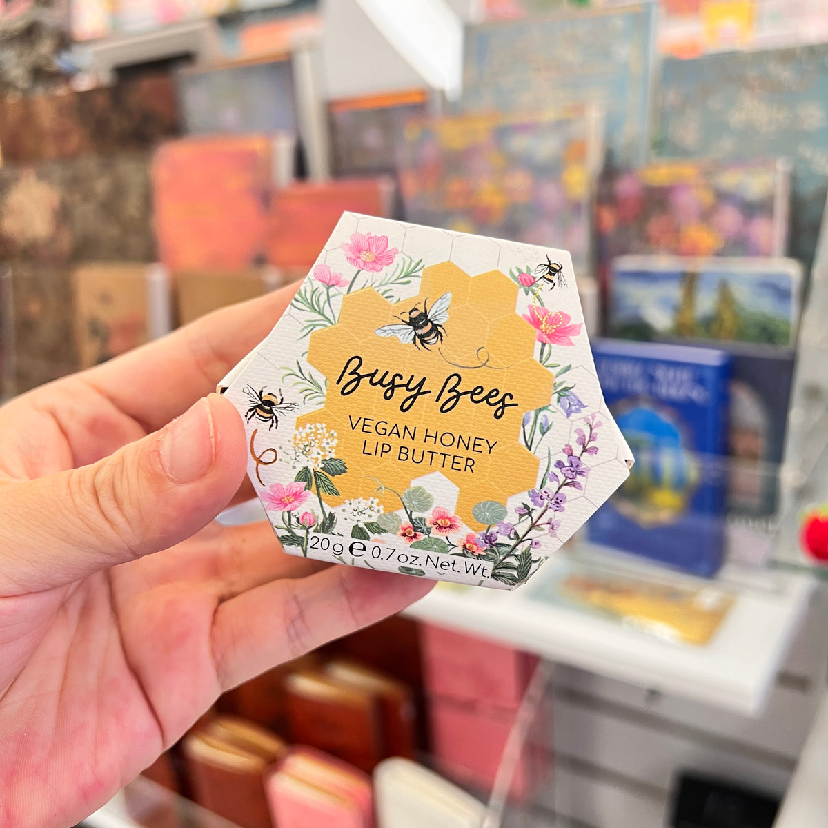 A hand holding a hexagonal box of 'Busy Bees Vegan Honey Lip Butter' in front of a blurred store background.