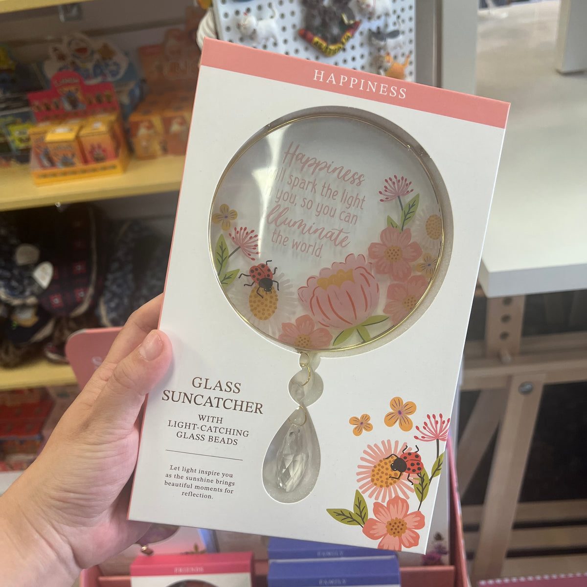 A hand holds a packaged glass suncatcher in a retail store setting. The product is a decorative glass mirror with floral and bee illustrations and the word 'HAPPINESS' at the top, with text reading 'GLASS SUNCATCHER' and 'WITH LIGHT CATCHING GLASS BEADS.