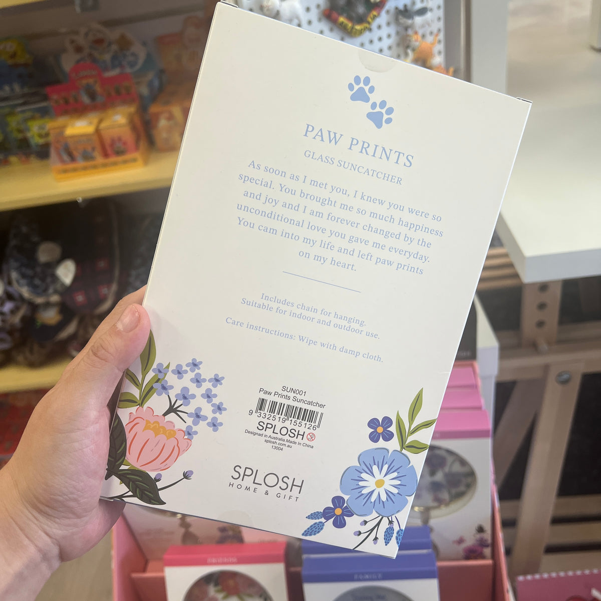 A hand holds the back of a SPLOSH PAW PRINTS glass suncatcher package in a retail store setting. The white card shows blue paw prints, floral illustrations, product text, and a barcode.
