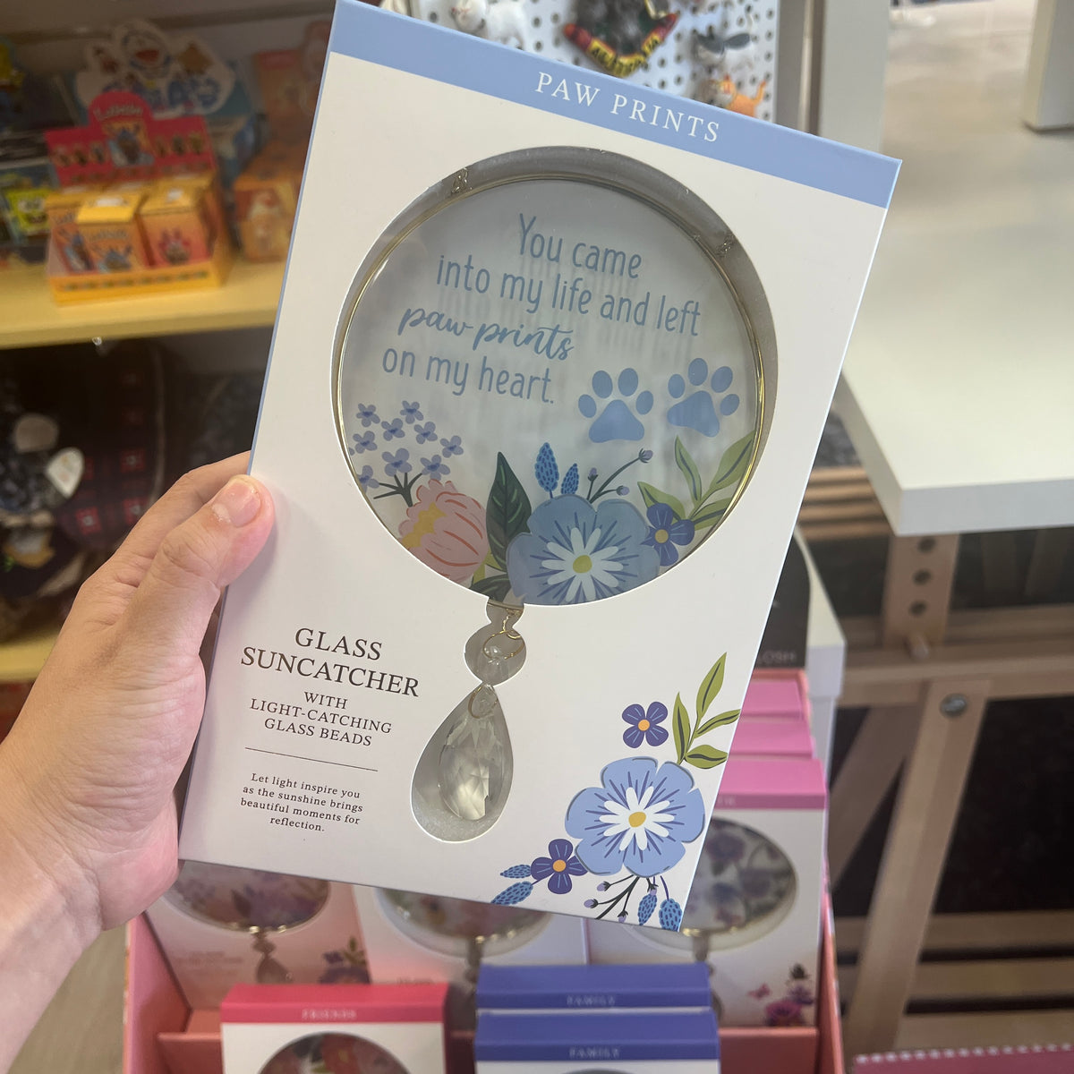 A hand holds a boxed glass suncatcher in a retail store setting. The package is white with blue 'PAW PRINTS' branding, floral illustrations, and a large printed depiction of the round glass suncatcher with paw prints and the phrase, 'You came into my life and left paw prints on my heart.