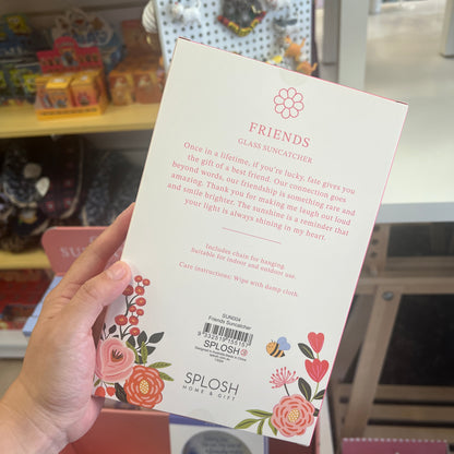 A hand holds the back of a SPLOSH Friends Glass Suncatcher box in a retail store setting. The packaging is white with pink text and colorful floral illustrations along the bottom.