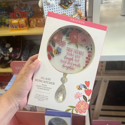 A hand holds a boxed glass sun catcher in a retail store setting. The package is white with pink floral artwork, a pink top band, and a clear teardrop-shaped glass catcher visible through a circular cutout.