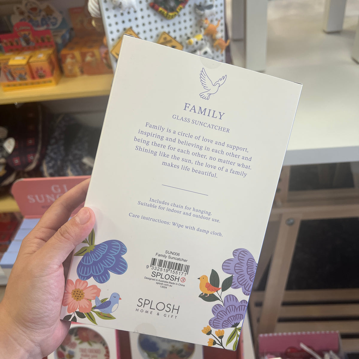 A hand holds a white product card for the back of a SPLOSH Family Glass Windcatcher, showing printed text, a barcode, and colorful floral and bird illustrations. The background shows a blurred retail store shelf with colorful items.