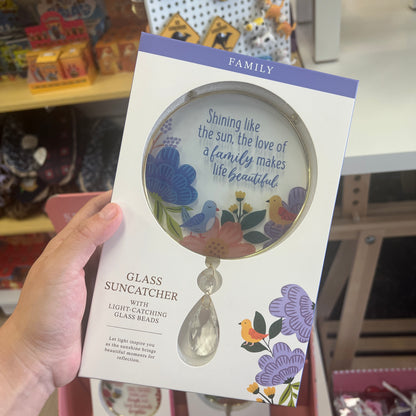 A hand holds a boxed glass suncatcher in a retail store setting. The package is white with purple accents, a large printed magnifying glass illustration, and the words 'FAMILY' and 'GLASS SUNCATCHER WITH LIGHT-CATCHING GLASS BEADS.